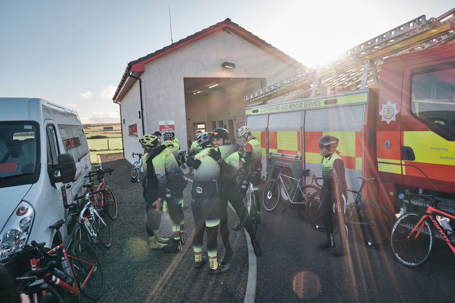Cyclists in green gear gather near a fire truck and van outside a rural fire station on a sunny day.