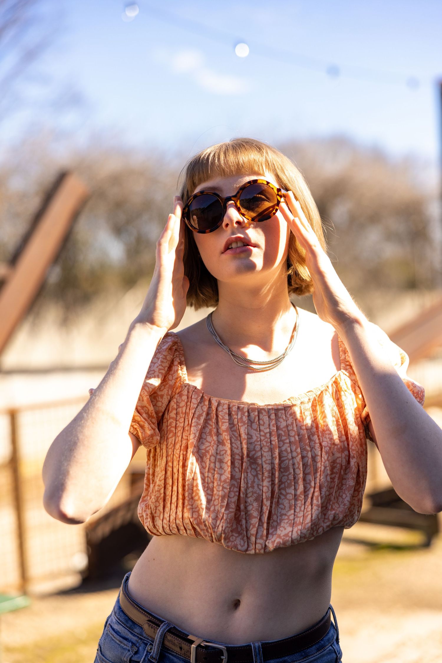 Young blonde woman in crop top and jeans adjusting tortoise shell sunglasses outdoors on a sunny day.