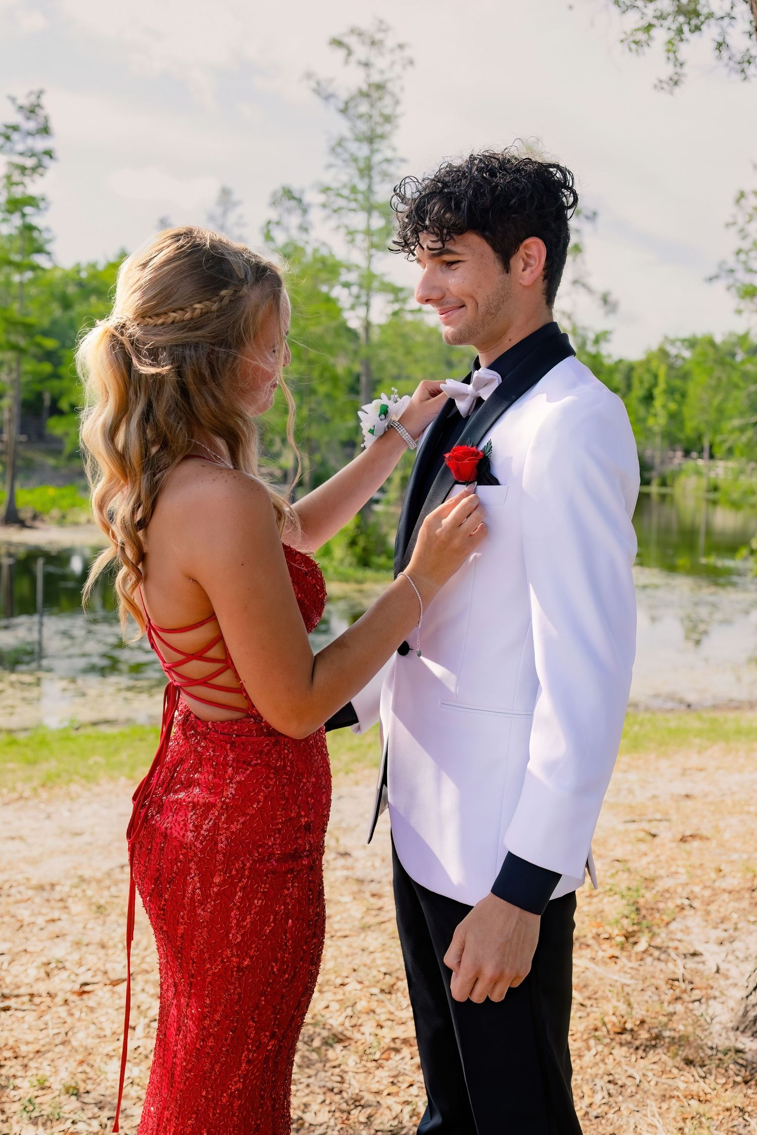 A smiling young man in a white tuxedo receives a boutonniere from a woman in a red sequin gown outdoors.