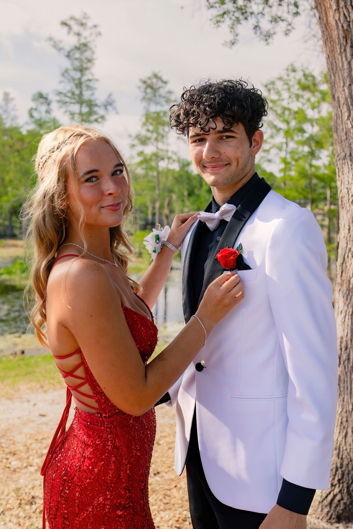 A teenage couple in prom attire, girl in red sequin gown pins boutonniere on boy in white tuxedo outdoors.