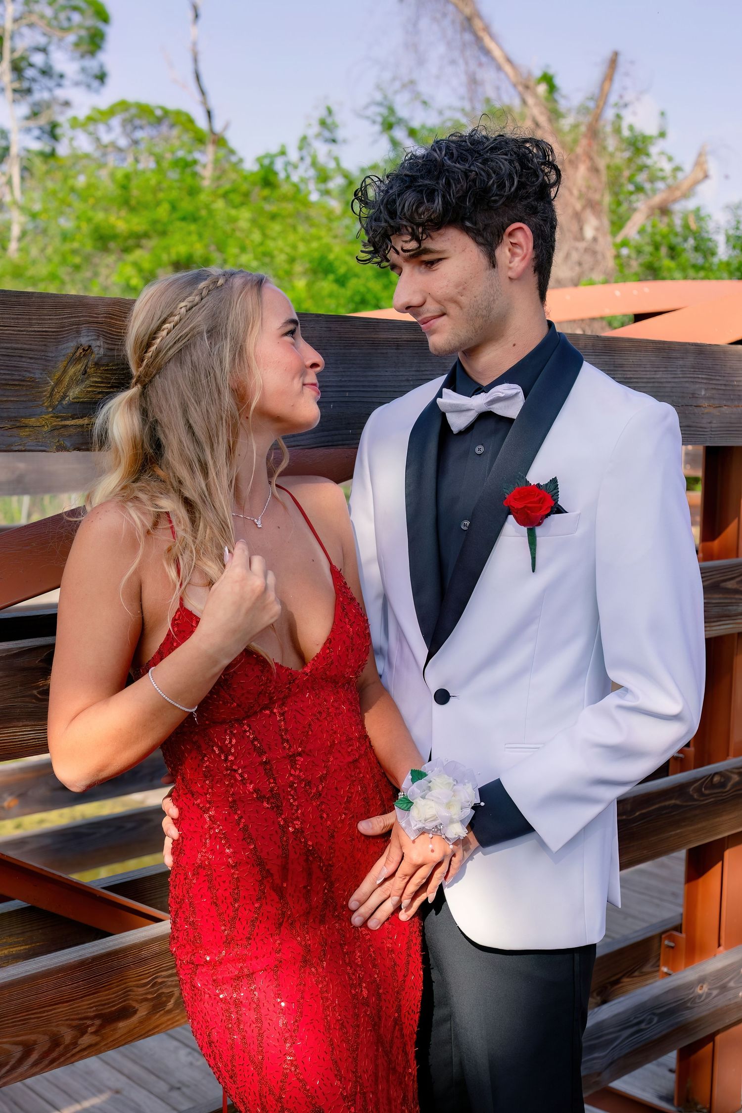 A couple dressed in prom attire, woman in red sequin gown and man in white tuxedo with red rose boutonniere, pose on wooden bridge.