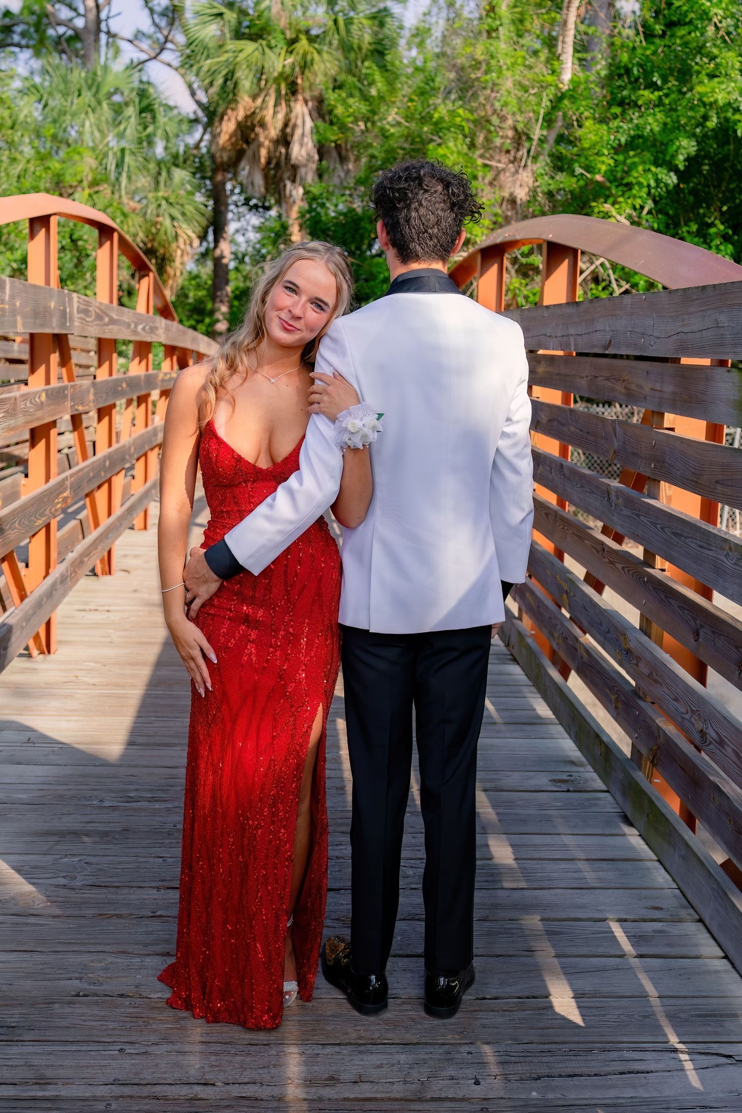 Couple in prom attire on wooden bridge, woman in red sequin gown, man in white tuxedo jacket.