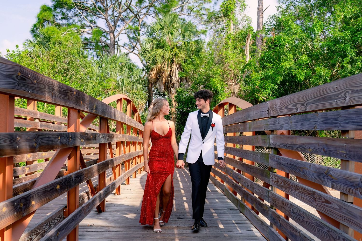 A couple in formal prom attire walks hand-in-hand on a wooden bridge surrounded by lush green trees.