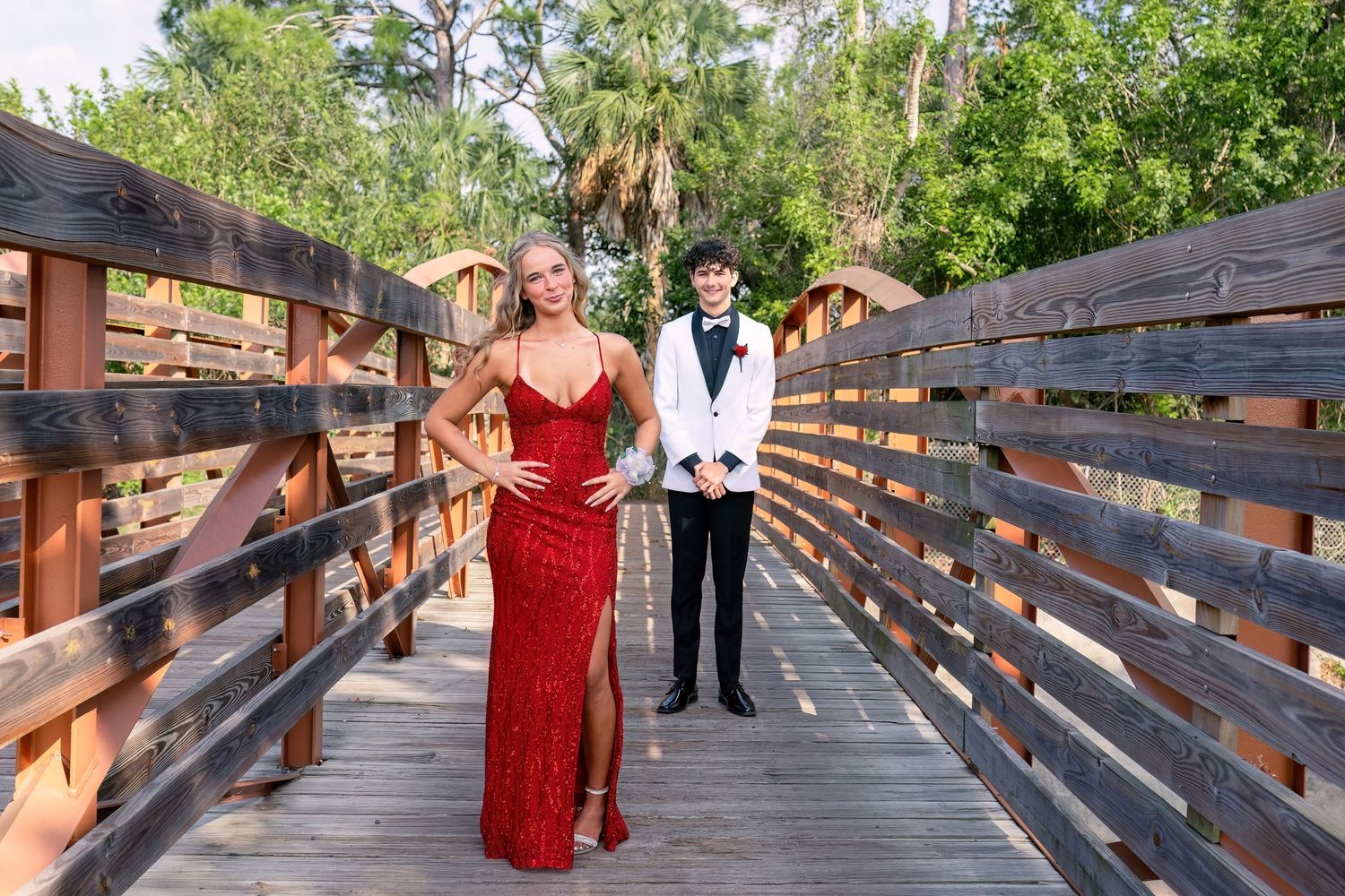A couple dressed in prom attire poses on a wooden bridge surrounded by lush greenery.