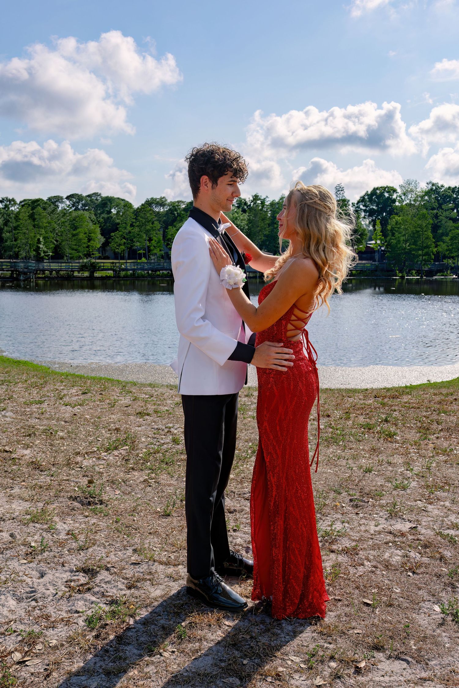 A prom couple in matching red dress and white tuxedo stand romantically by a lake on a sunny day.