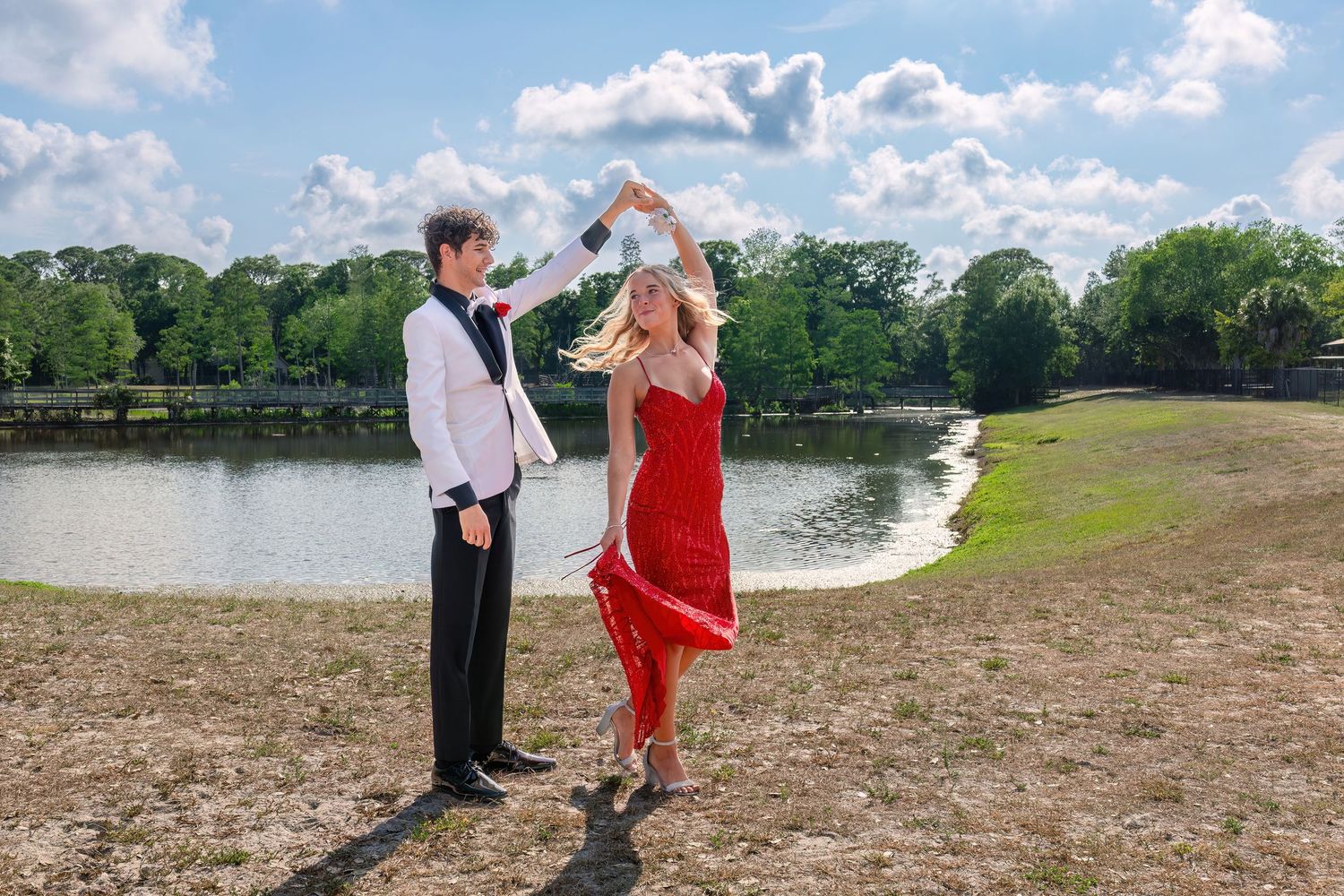 A couple in formal attire dances outdoors near a lake, the woman in a red dress being twirled by her partner.