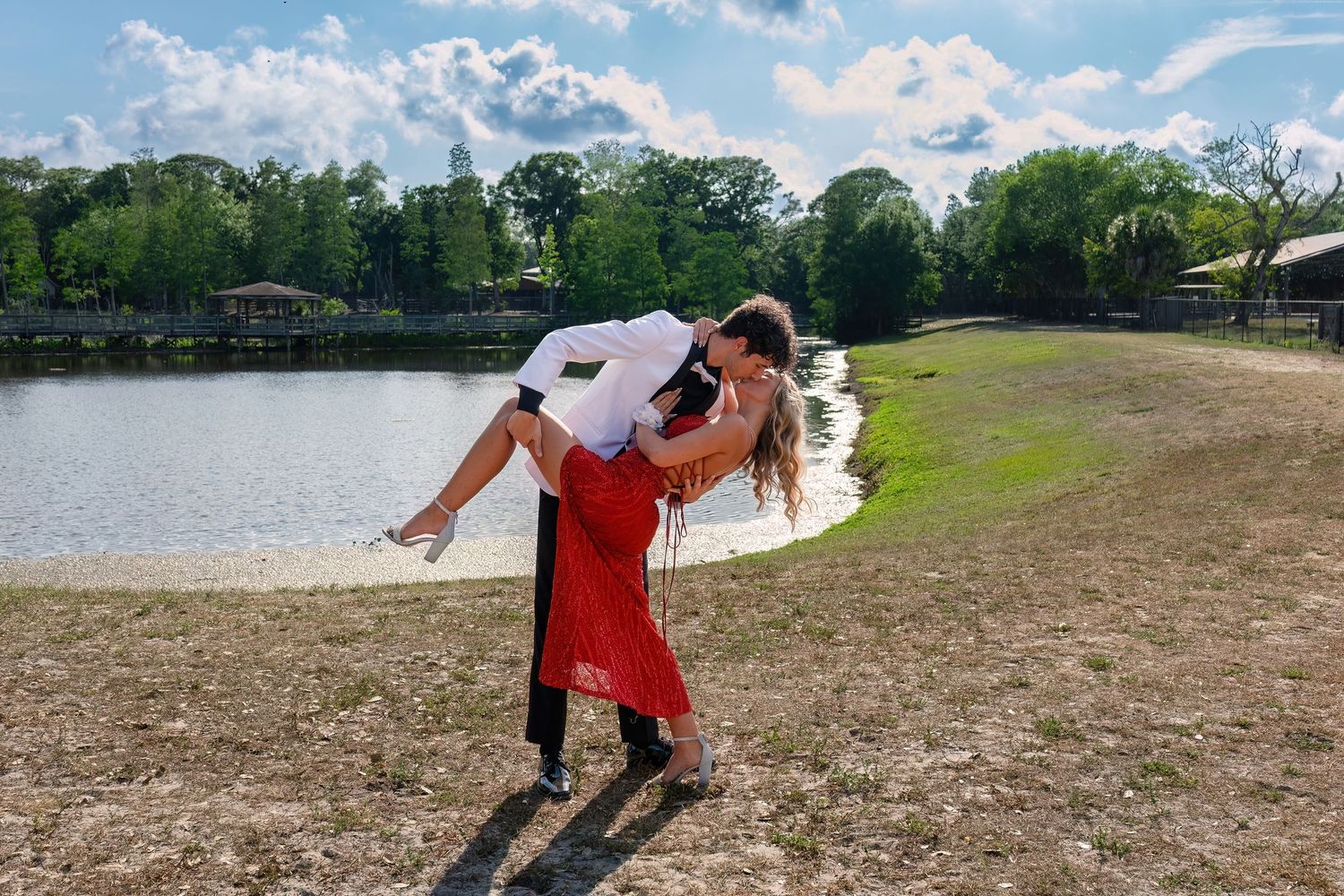 Couple in formal wear sharing romantic dip pose by a lake, man in white shirt holding woman in red dress outdoors.