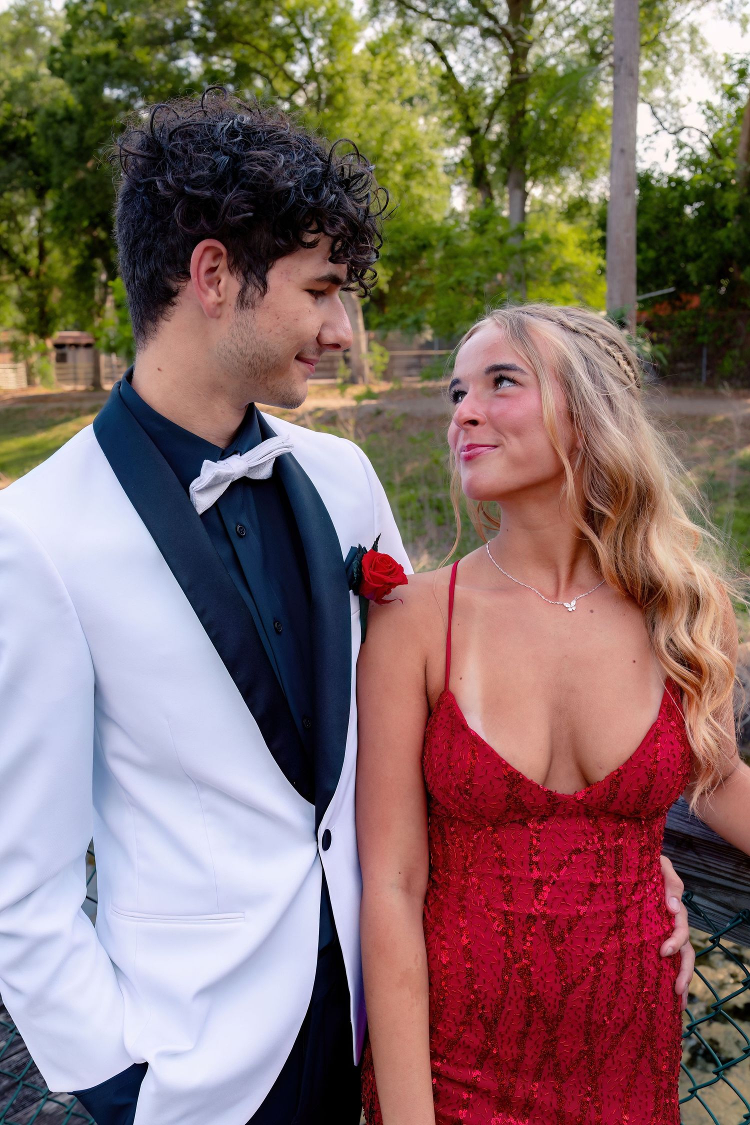 A young couple dressed in prom attire, man in white tuxedo with red boutonniere, woman in red sequin gown, smiling at each other.