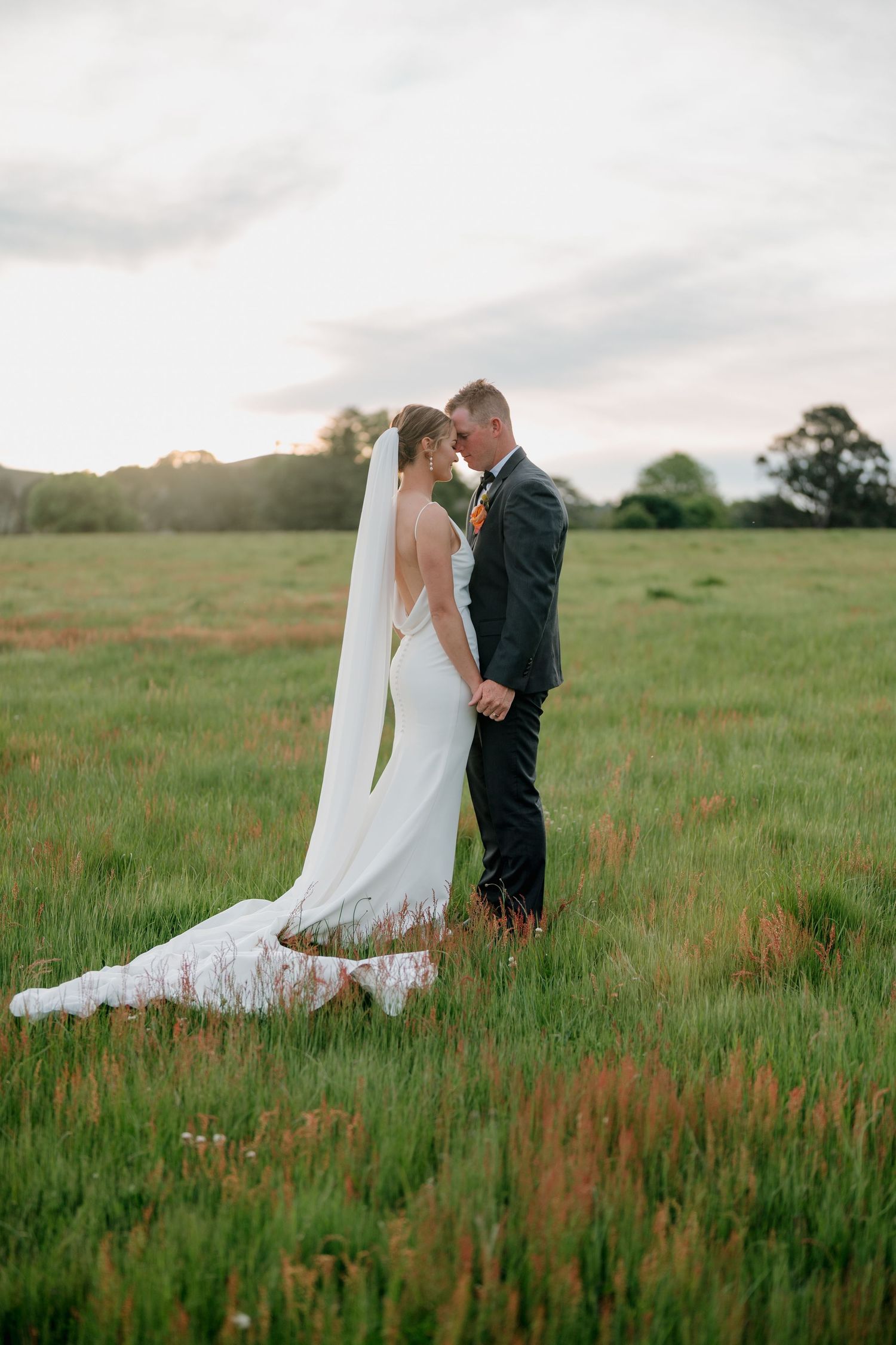 Bride and groom share a romantic moment in a lush green field at sunset, bride wearing a flowing white gown with veil.