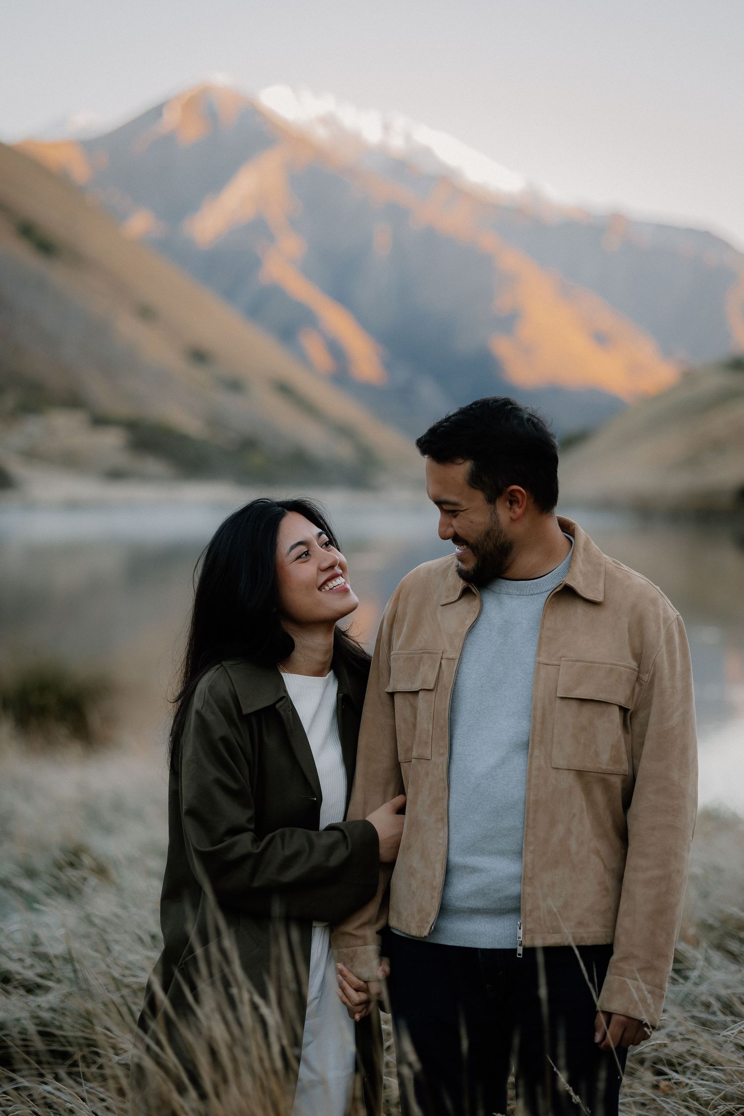 Couple smiling at each other near a mountain lake at sunset, woman in olive jacket, man in tan suede shirt.