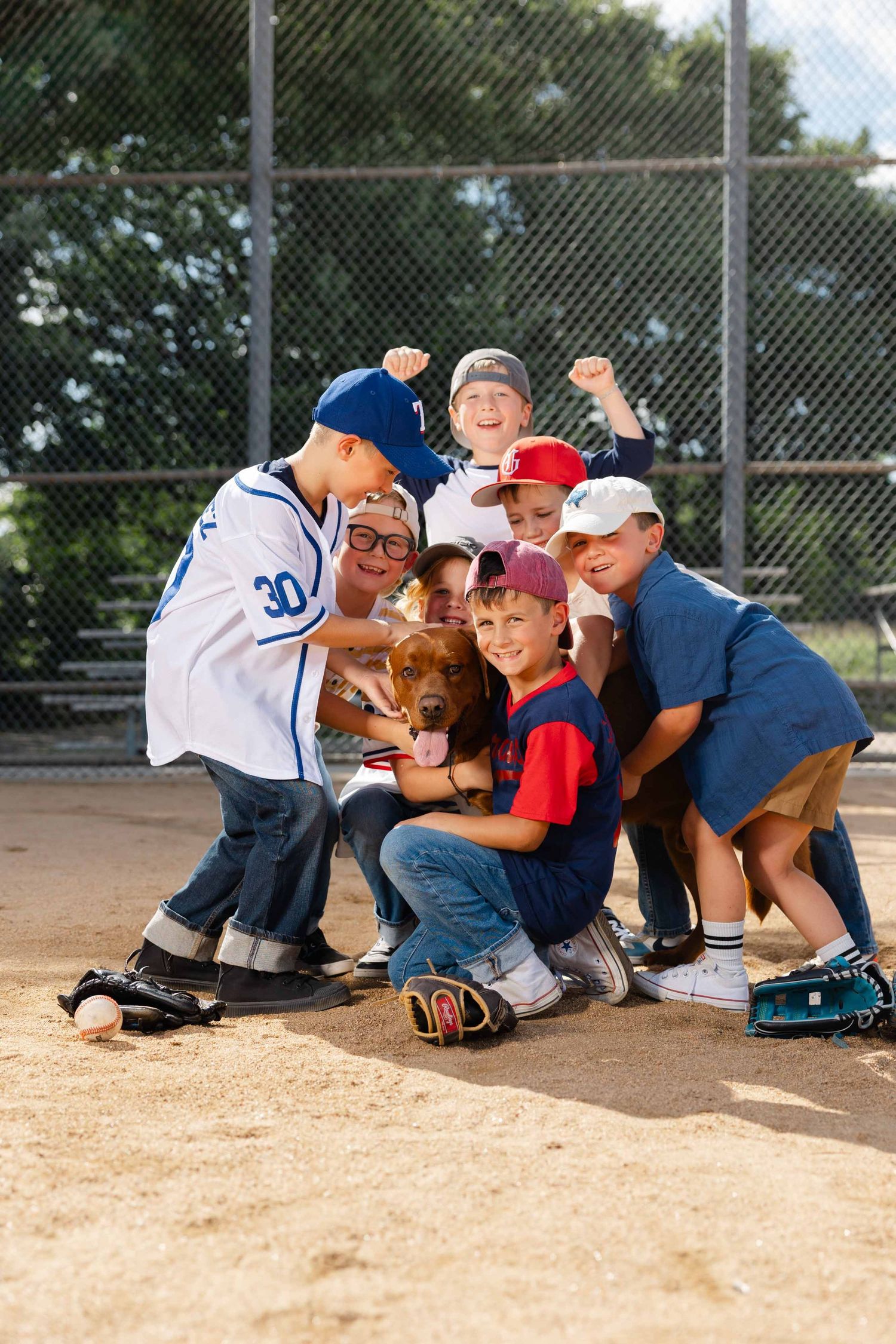 Youth baseball team huddles together with a brown dog on a dirt field, smiling and celebrating after a game.