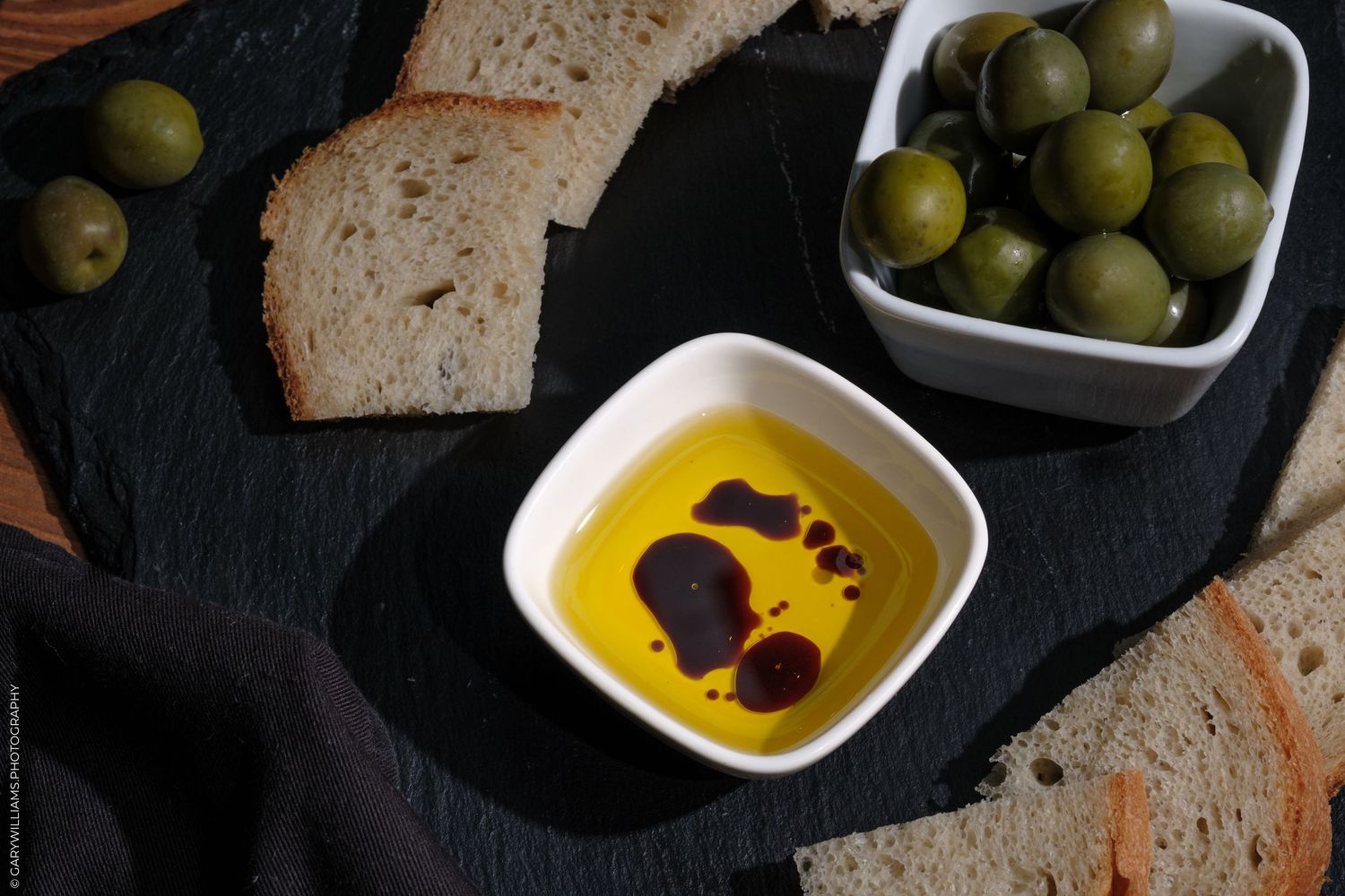 Olive oil dip with balsamic in white bowl, green olives, and sliced bread on dark slate board.