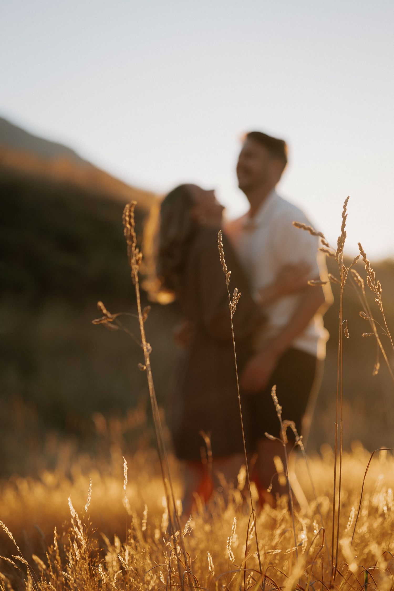 Couple sharing a romantic kiss at golden hour in a sunlit field with warm lens flare and rolling hills.