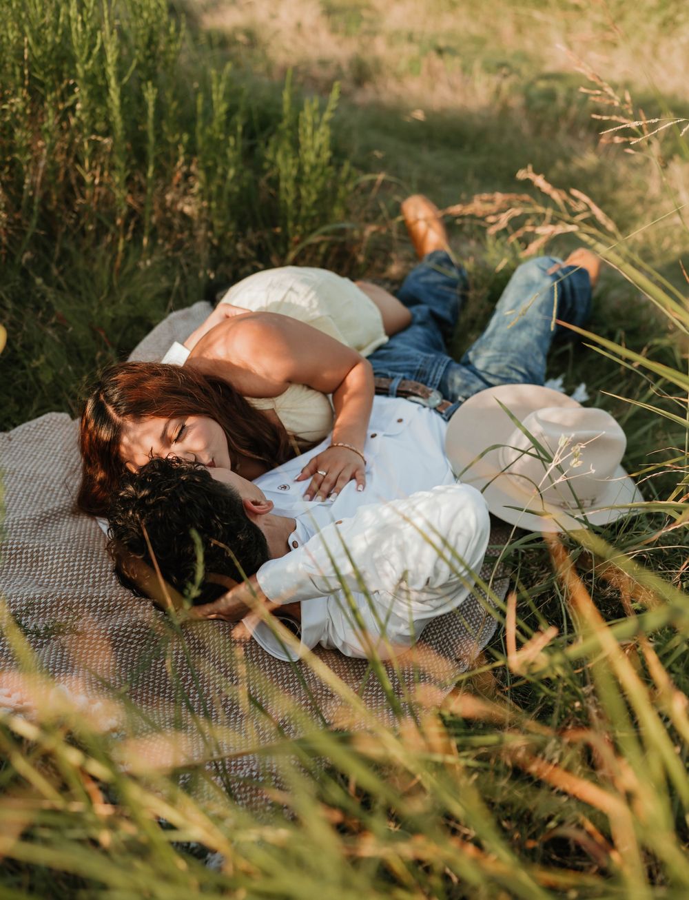Couple lying on a blanket in a grassy field, sharing a romantic moment in warm golden sunlight.