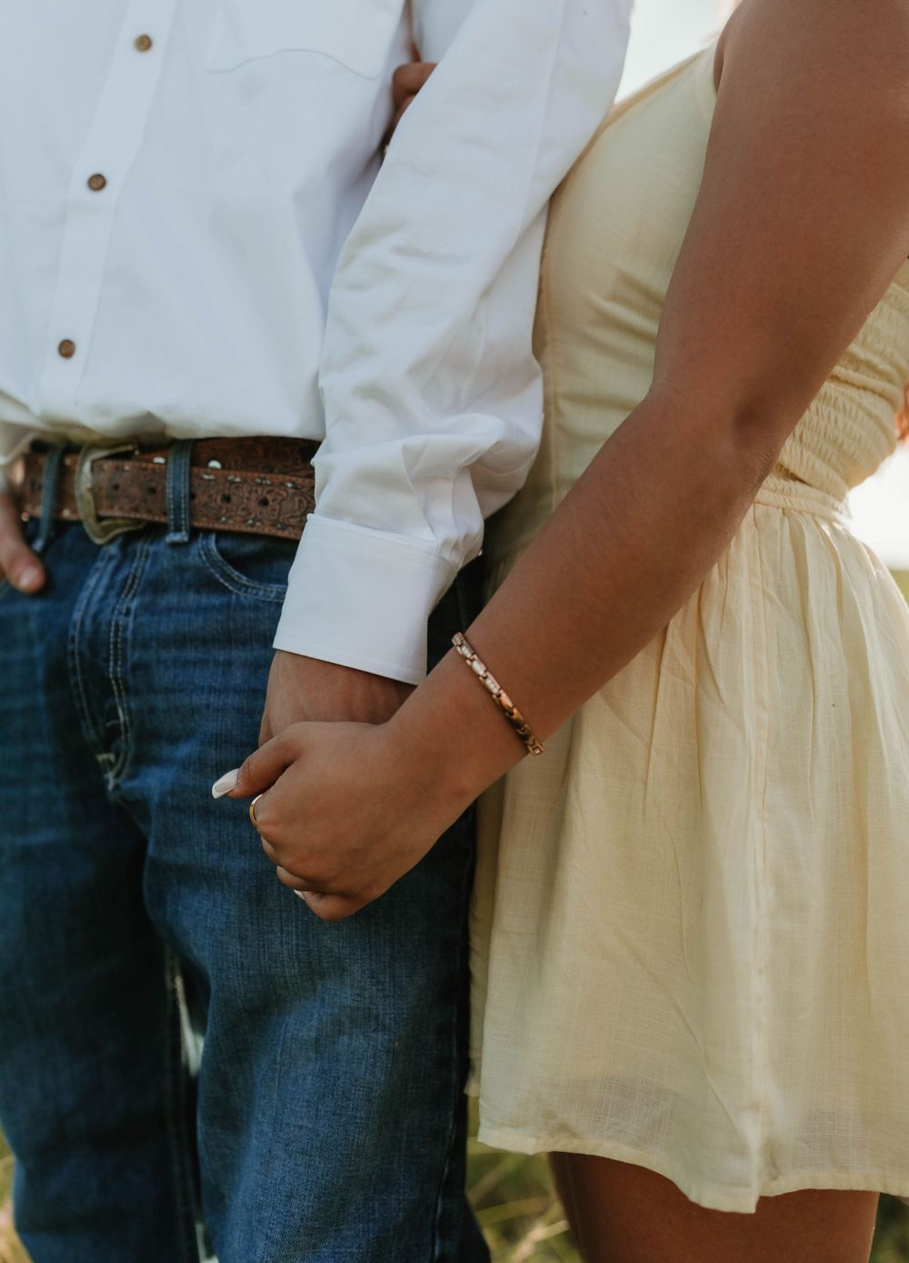 Couple holding hands, man in white shirt and jeans with brown belt, woman in cream dress with gold bracelet.