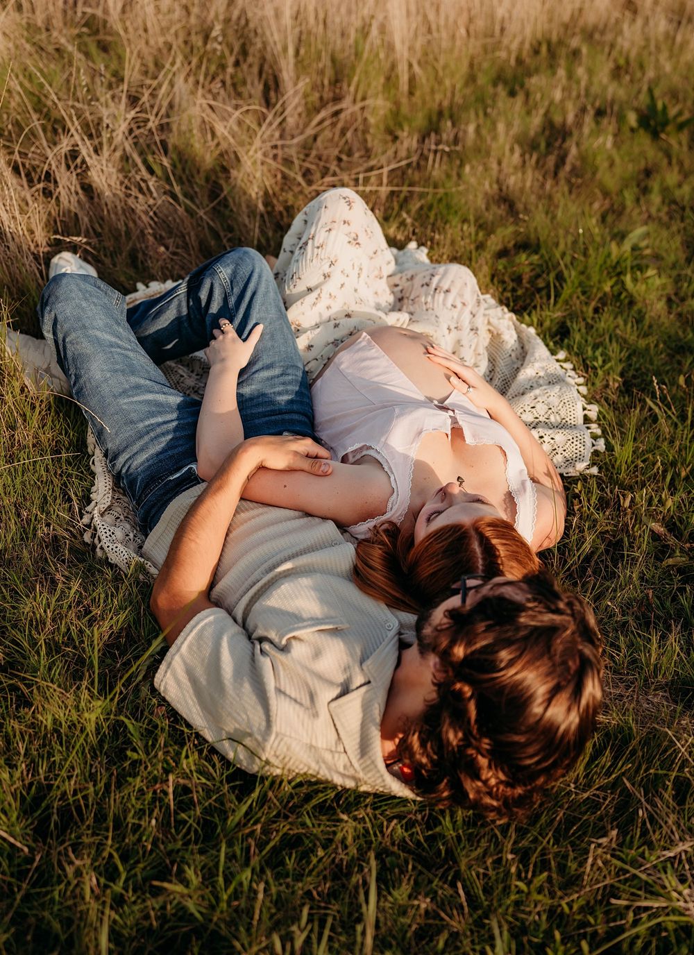 Couple lying in a grassy field at sunset, man resting head on pregnant woman's belly.