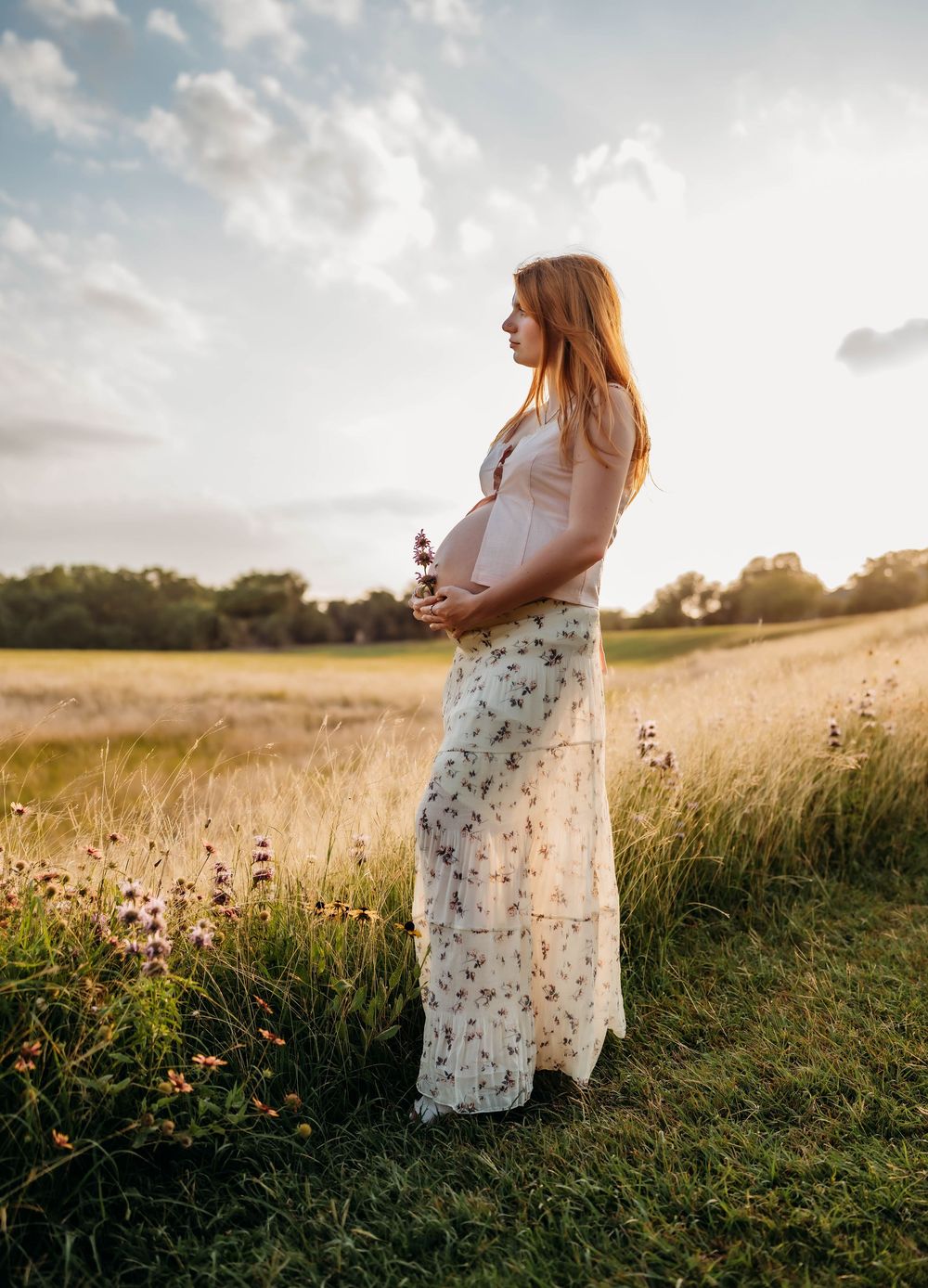 Pregnant woman in white floral maxi dress cradles baby bump while standing in a sunlit meadow at sunset.