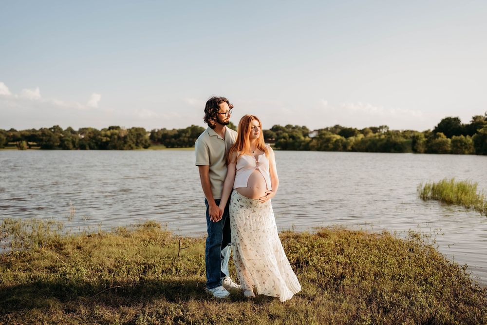 Couple holding hands by a lake at golden hour, pregnant woman in floral dress, man in casual attire.