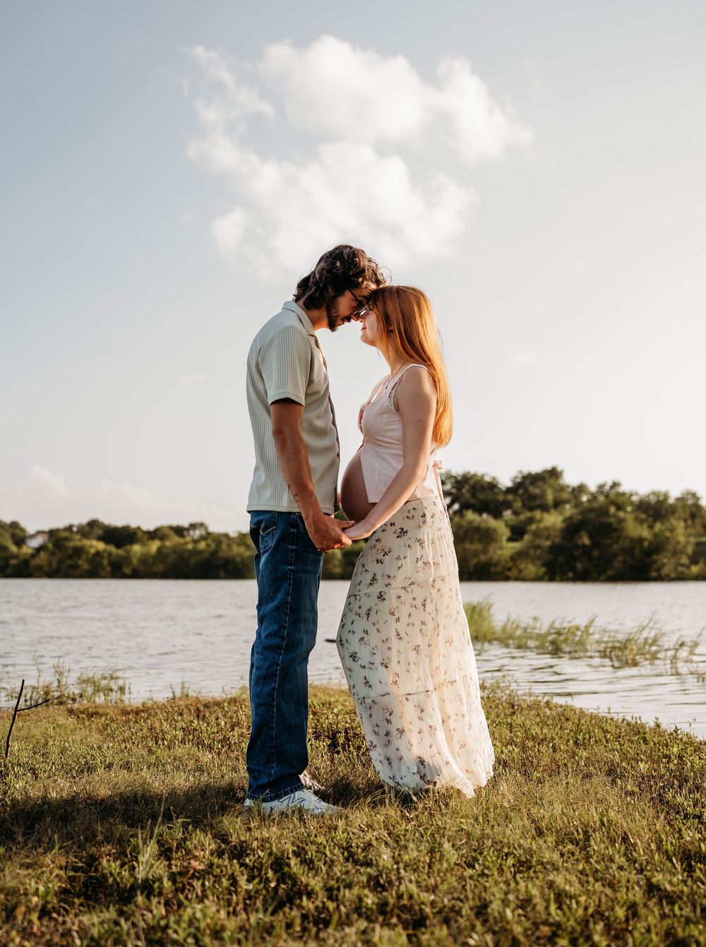Expecting couple holding hands and touching foreheads by a lake under a blue sky.