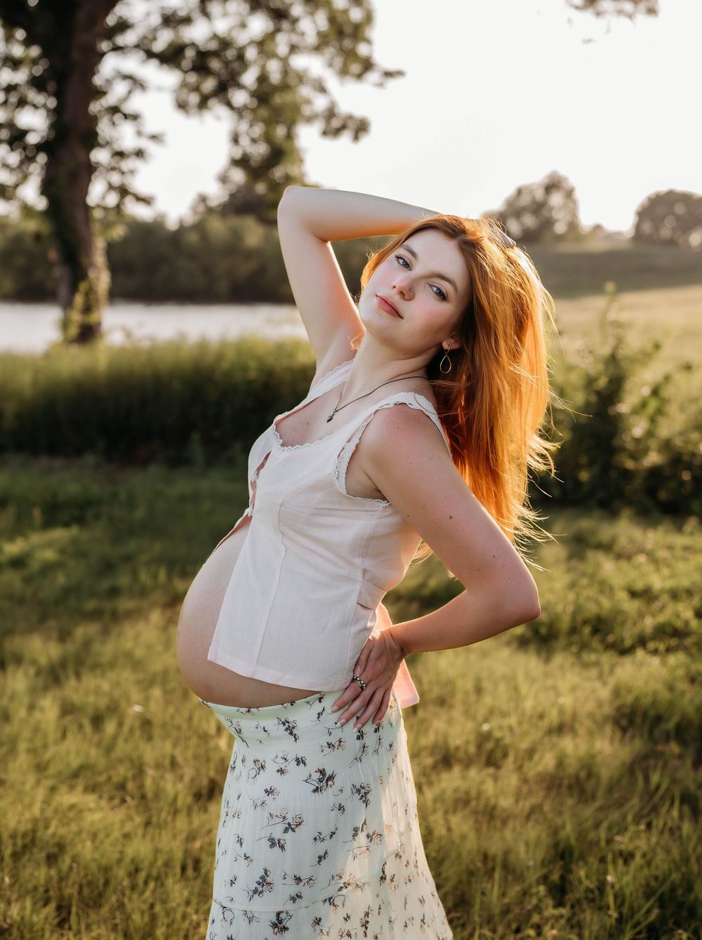 Pregnant redhead woman poses in white floral skirt and crop top during golden hour in a lush green field.