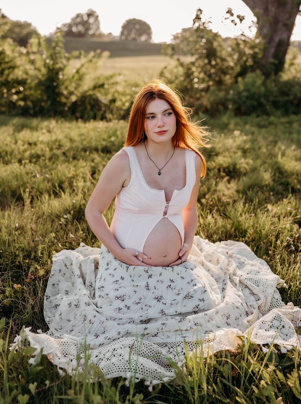 Pregnant redhead woman sitting in a sunlit meadow, wearing a white crop top and floral skirt.