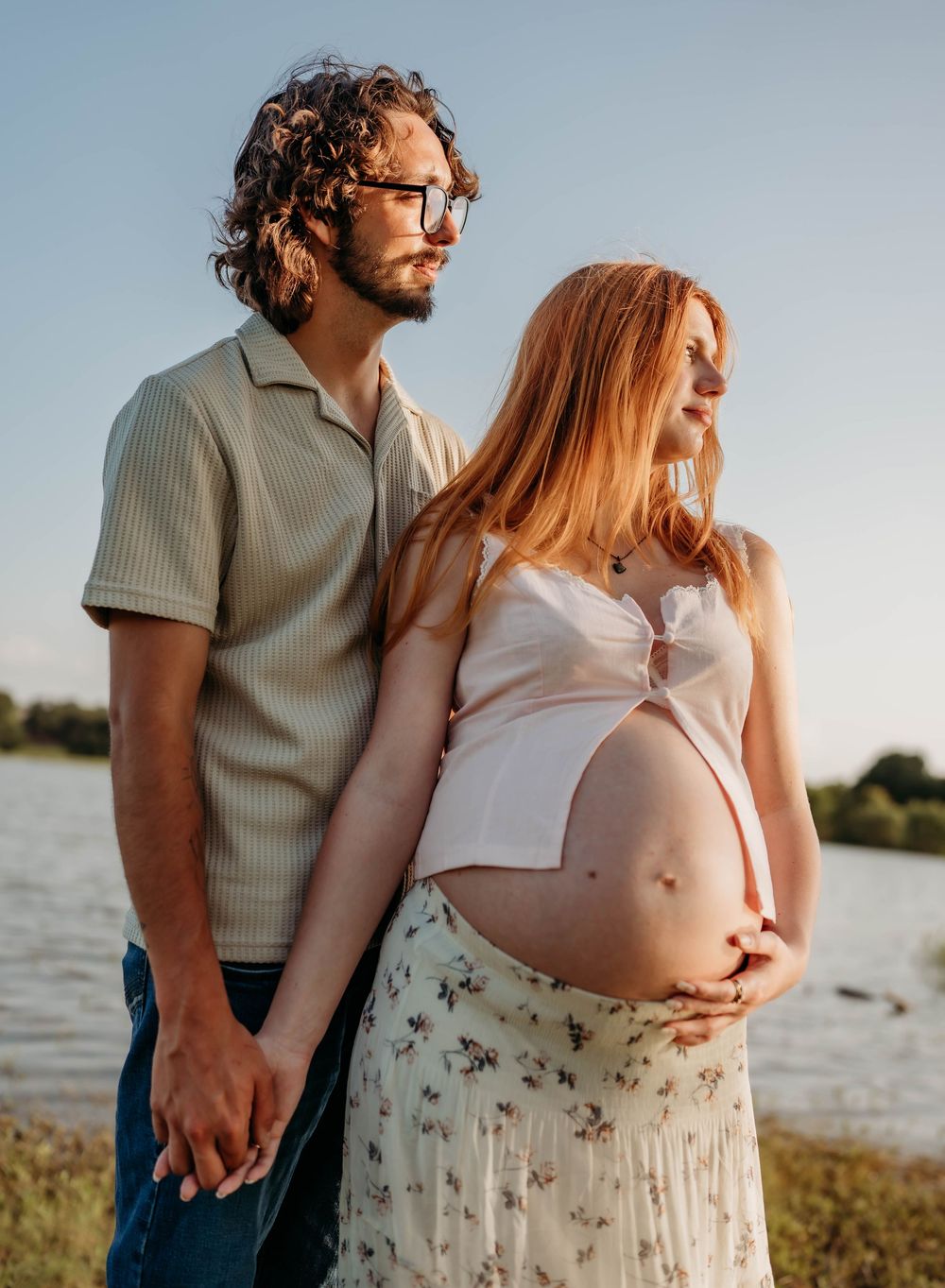 Pregnant couple holds hands by a lake at sunset, woman in floral skirt cradling her baby bump.