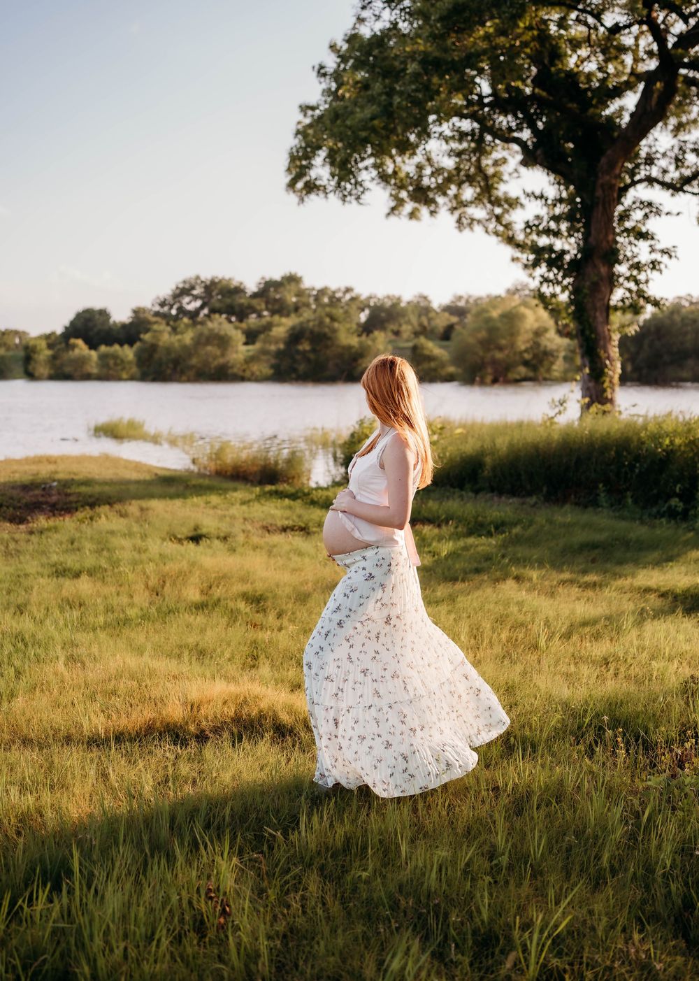 Pregnant woman in floral maxi skirt standing by a sunlit lake surrounded by lush green grass and trees.