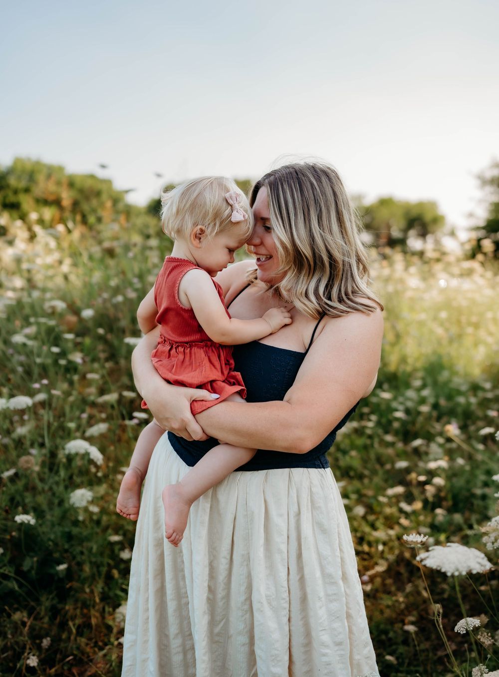 A mother holds her toddler in a sunlit wildflower field, both sharing a tender nose-to-nose moment.
