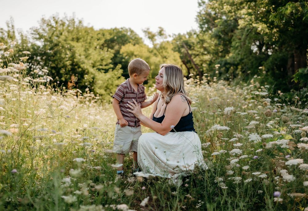 A mother and young son share a tender moment in a sunlit wildflower meadow surrounded by lush green trees.