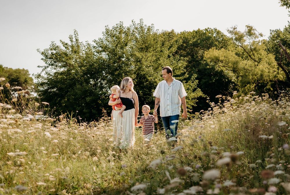 Family of four walking through a sunny wildflower meadow with lush green trees in the background.