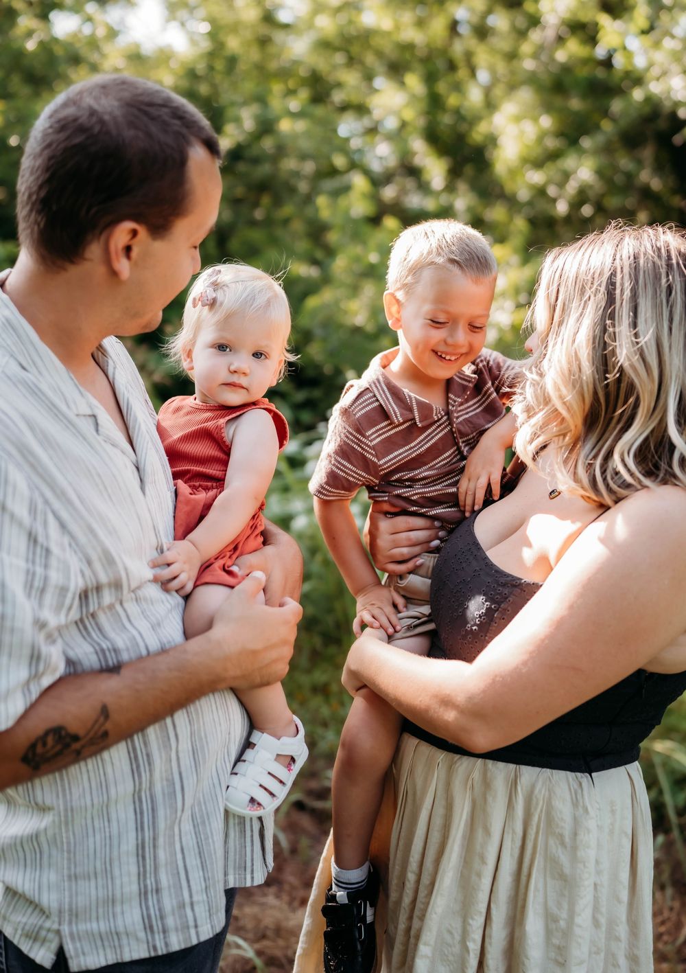 A young family with two toddlers enjoying a sunny outdoor moment, parents holding children while smiling in a lush green setting.
