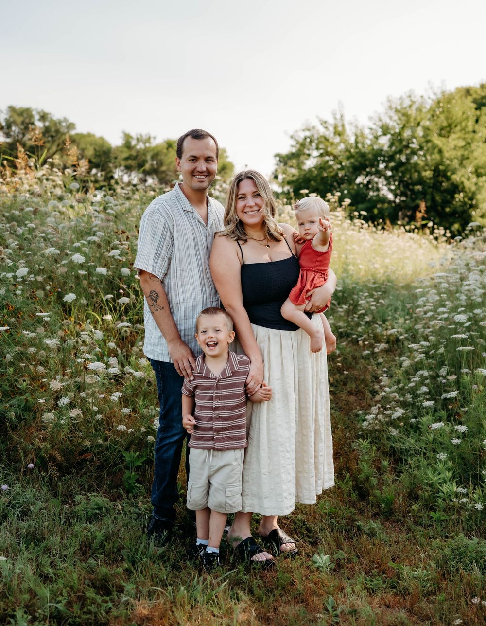 Family of four smiling in a wildflower field at golden hour, parents holding young children outdoors.