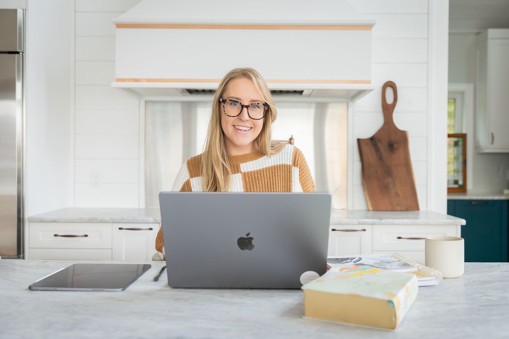 Smiling woman with glasses working on MacBook laptop at white kitchen counter with books and tablet nearby.