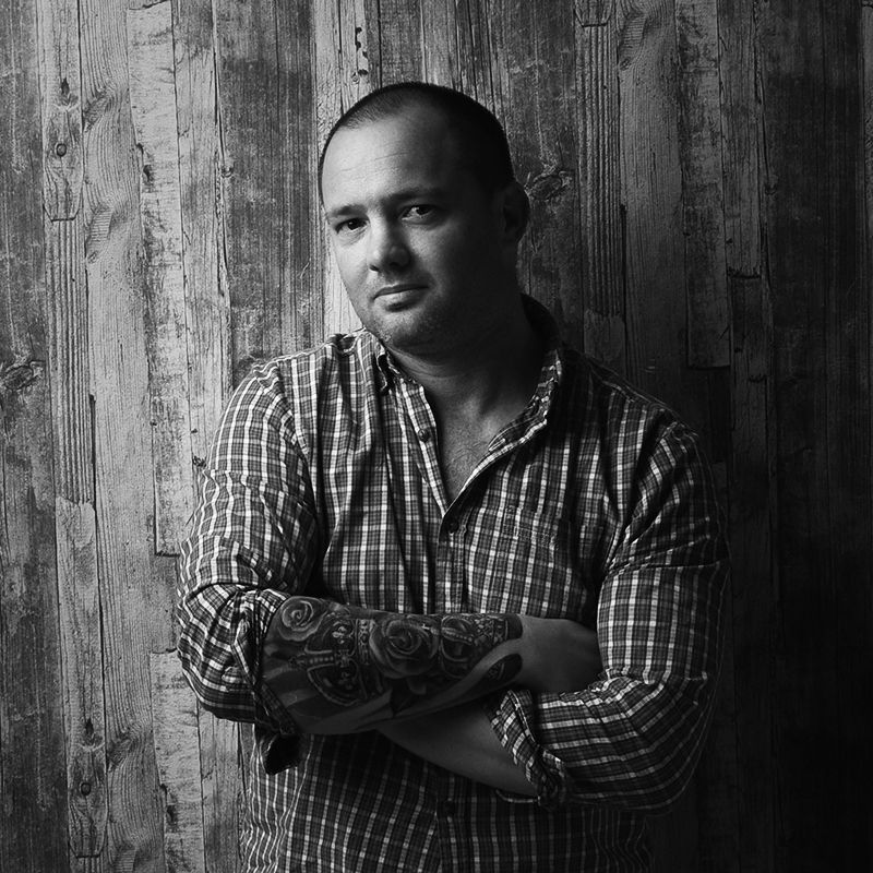 Tattooed man with crossed arms in plaid shirt leaning against rustic wood wall, serious expression.
