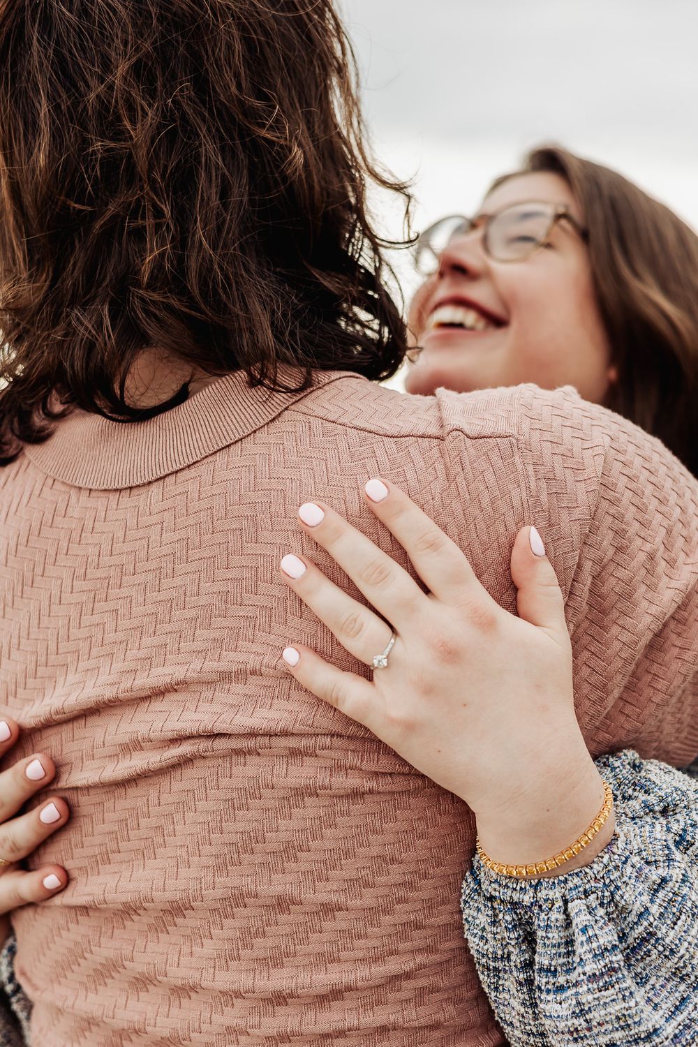 Two women embracing and laughing, one displaying an engagement ring and gold bracelet, wearing pink sweaters outdoors.