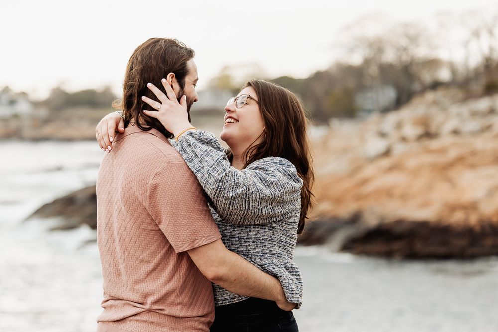 A couple embraces and smiles at each other on a rocky beach, sharing a romantic moment outdoors.