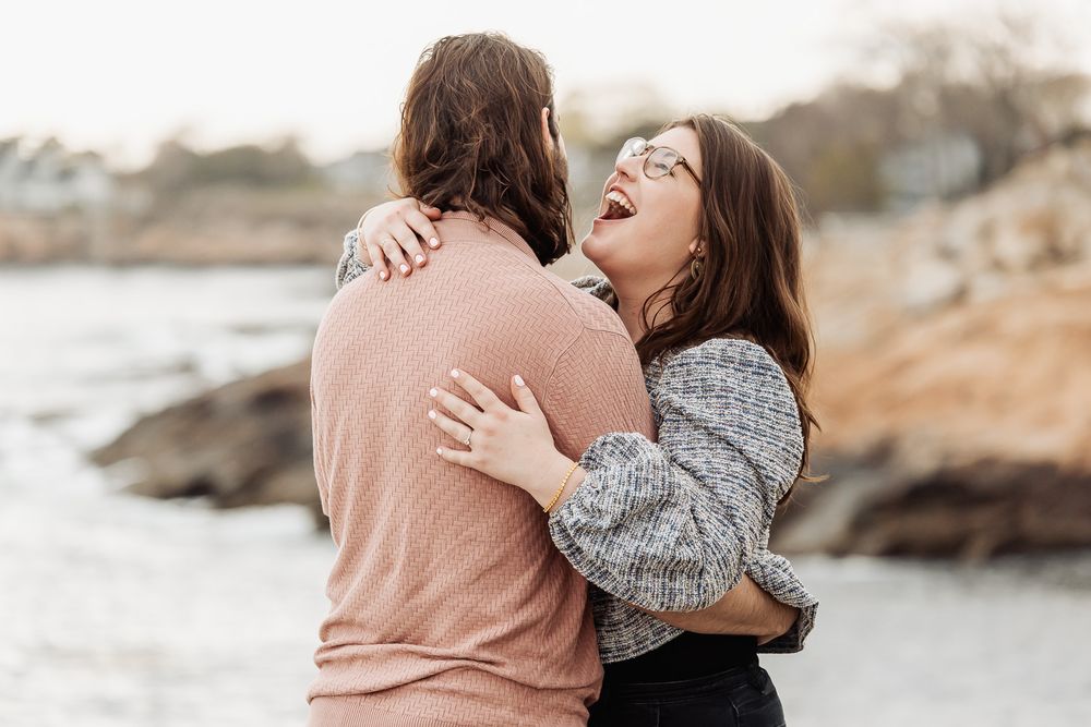A joyful couple embraces and laughs together on a rocky beach shore during an outdoor engagement photo session.