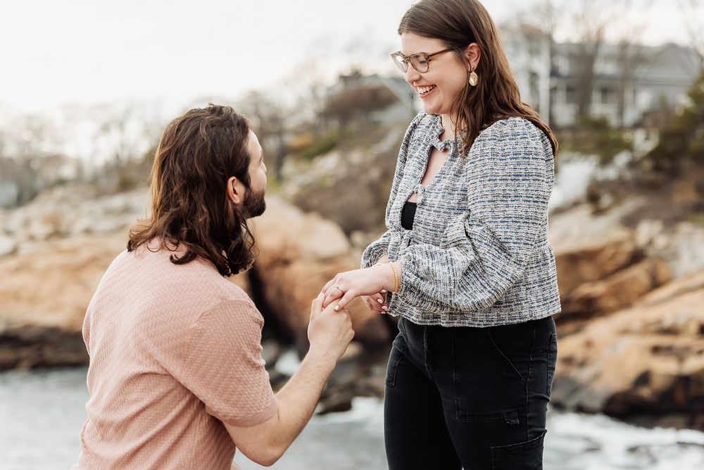 Man kneeling to propose to smiling woman near rocky ocean shore.