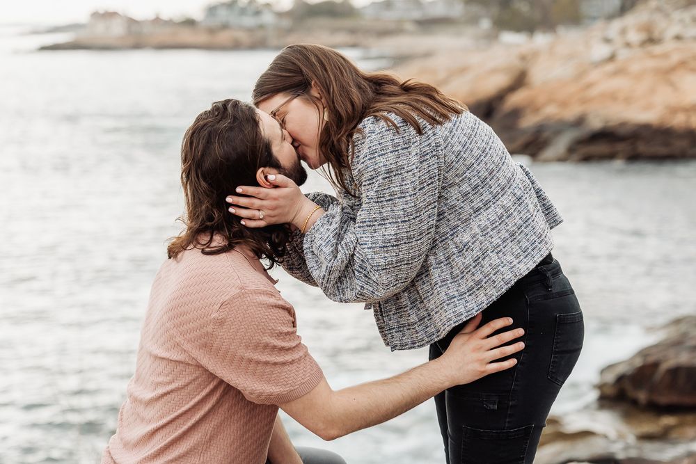 Two women sharing a tender moment by rocky seaside, one kneeling as they touch foreheads together affectionately.