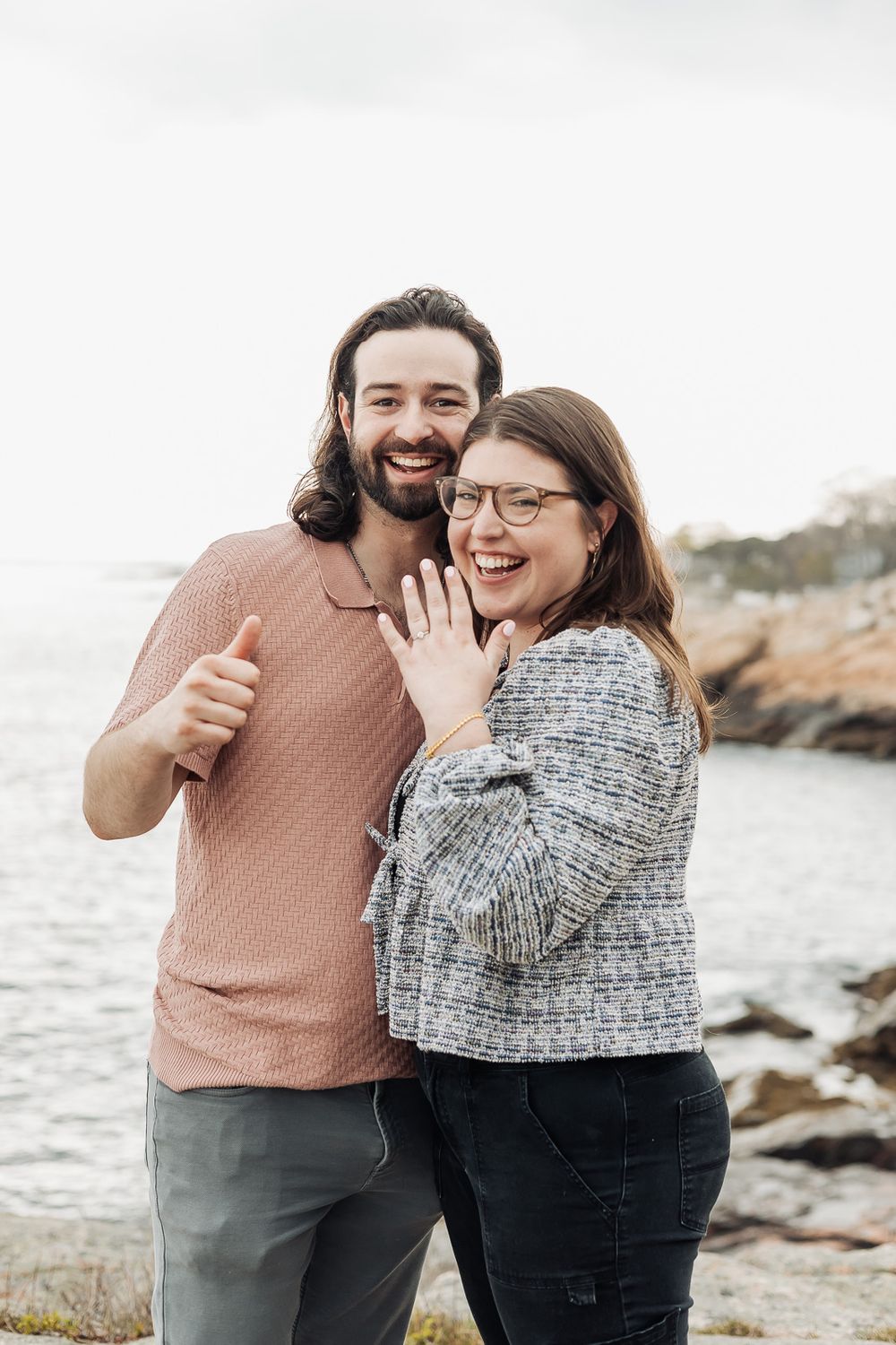 Joyful engaged couple showing off ring near rocky ocean coastline.