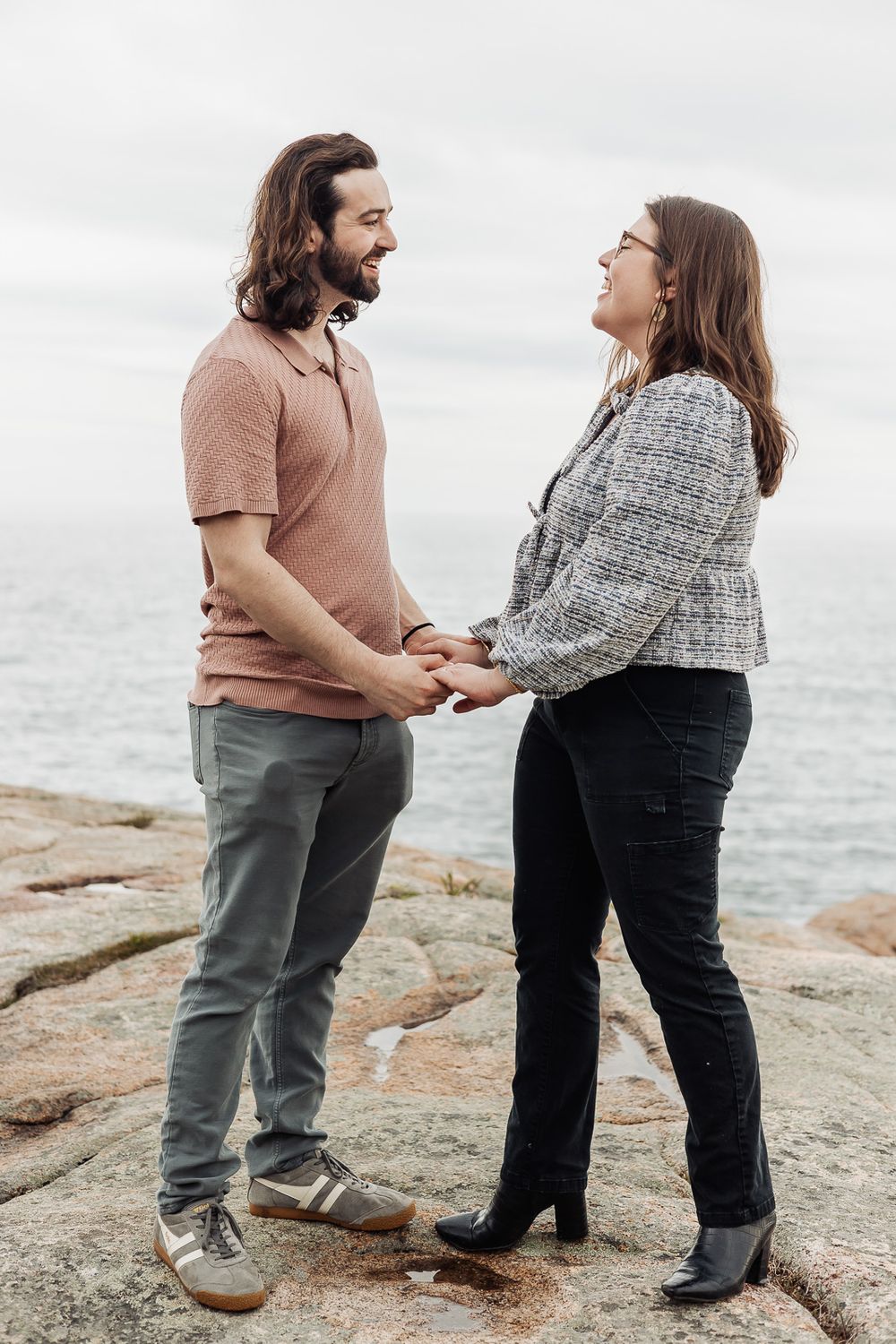 A couple holds hands and smiles at each other on rocky coastal cliffs with ocean views behind them.