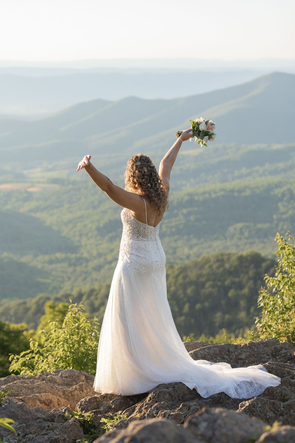 Bride in white lace gown raises bouquet triumphantly on rocky mountain overlook with scenic valley views.