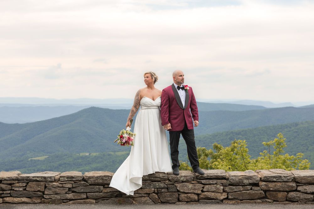 Bride and groom pose before stunning Blue Ridge Mountain panorama, she in white gown, he in burgundy tuxedo.