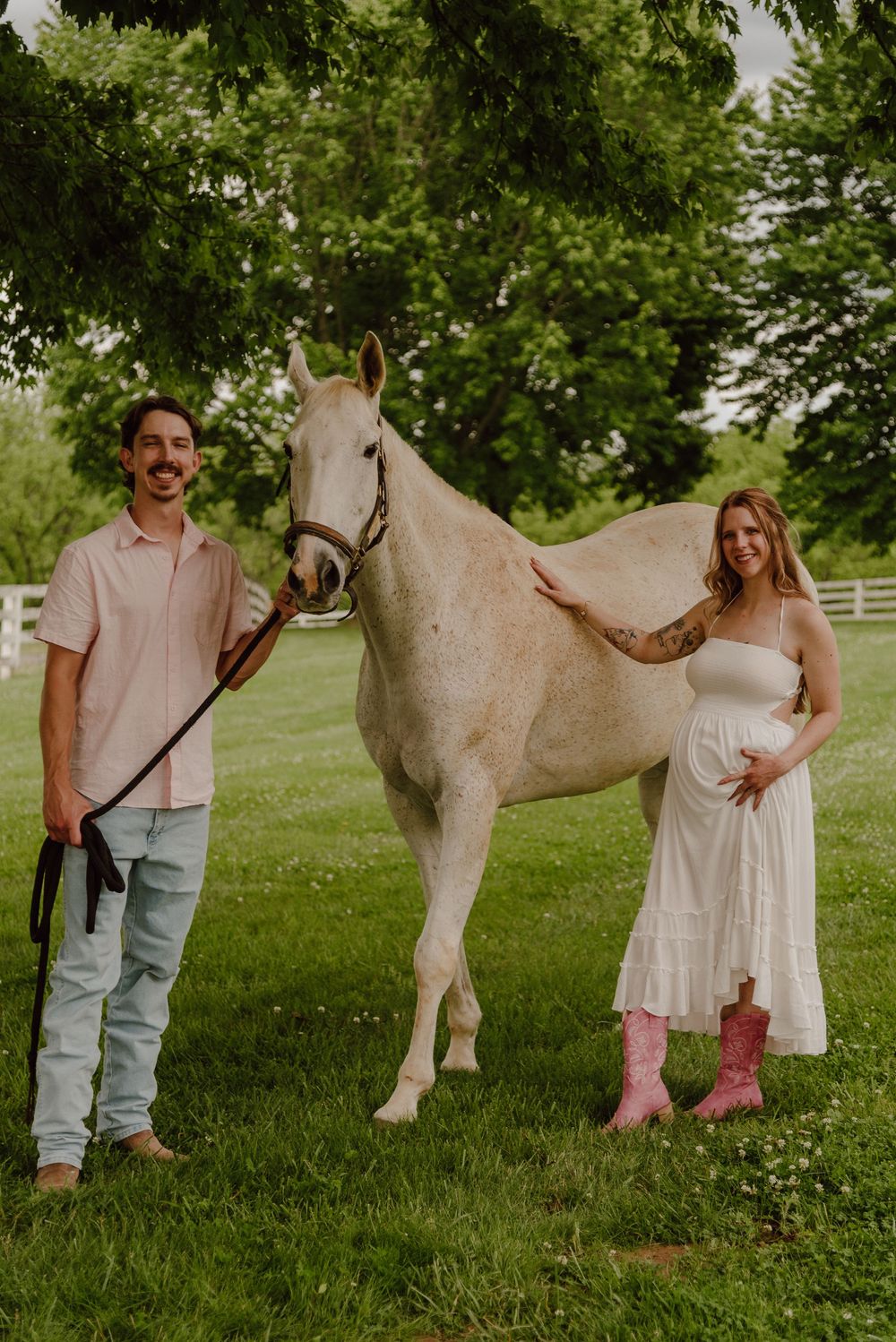Pregnant couple in pink and white outfits standing beside a white horse on a green lawn.