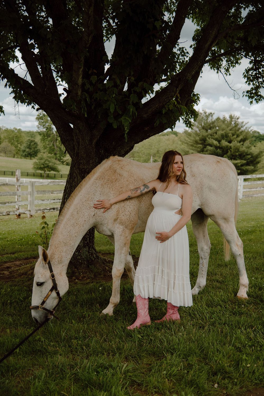 Pregnant woman in white dress and pink boots standing beside a white horse under a tree.