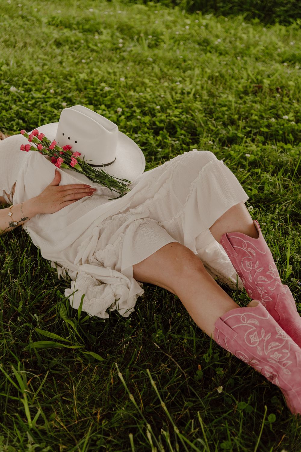 Woman in white dress and pink cowboy boots lying on green grass, holding a floral white cowboy hat.