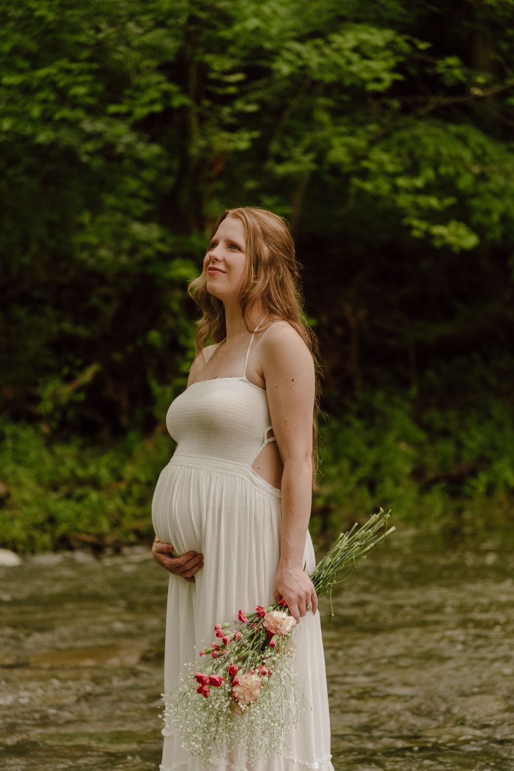 Pregnant woman in white dress holding flowers by a creek, surrounded by lush green trees.