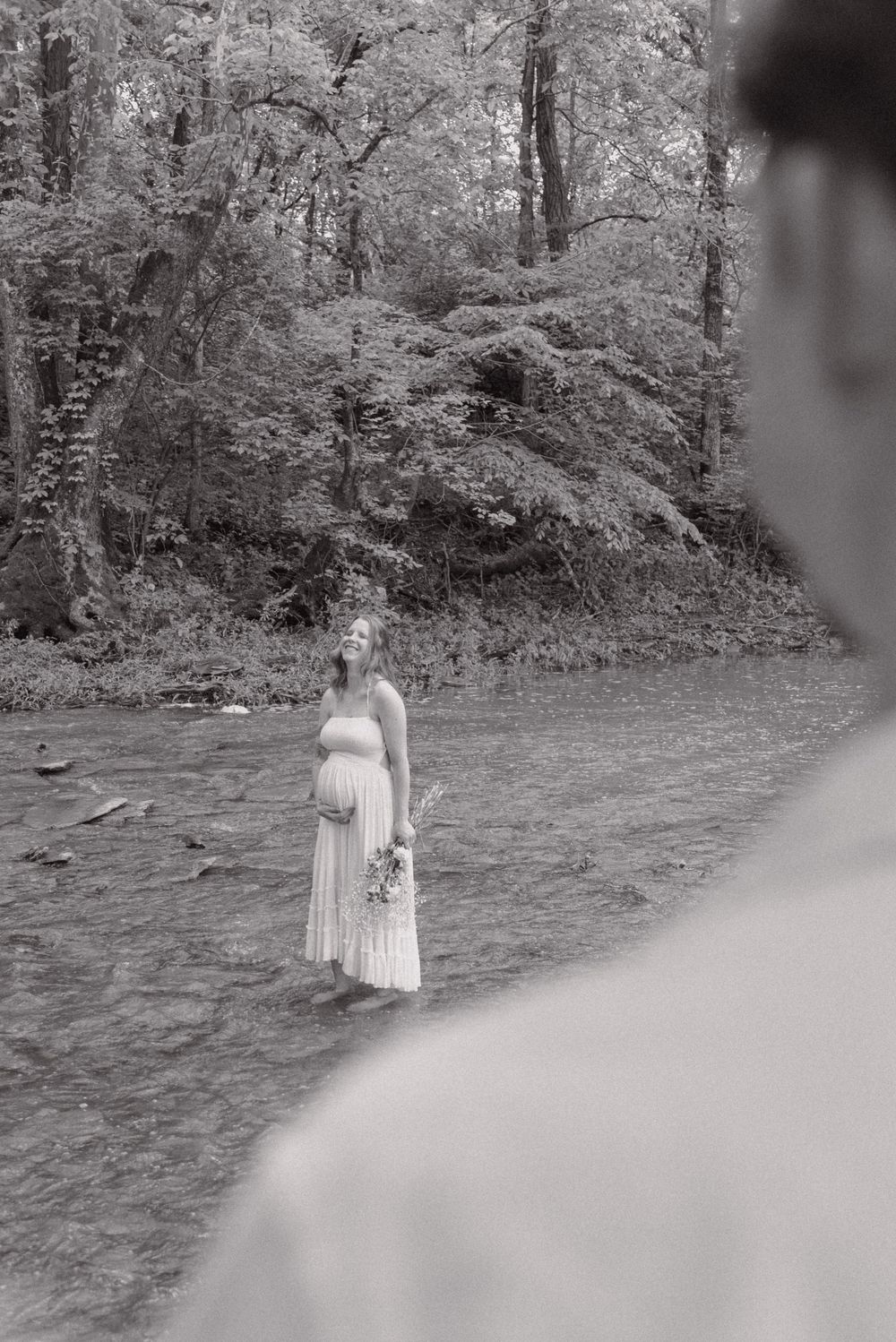 Black and white photo of a pregnant woman in a white dress standing in a shallow creek surrounded by lush forest trees.