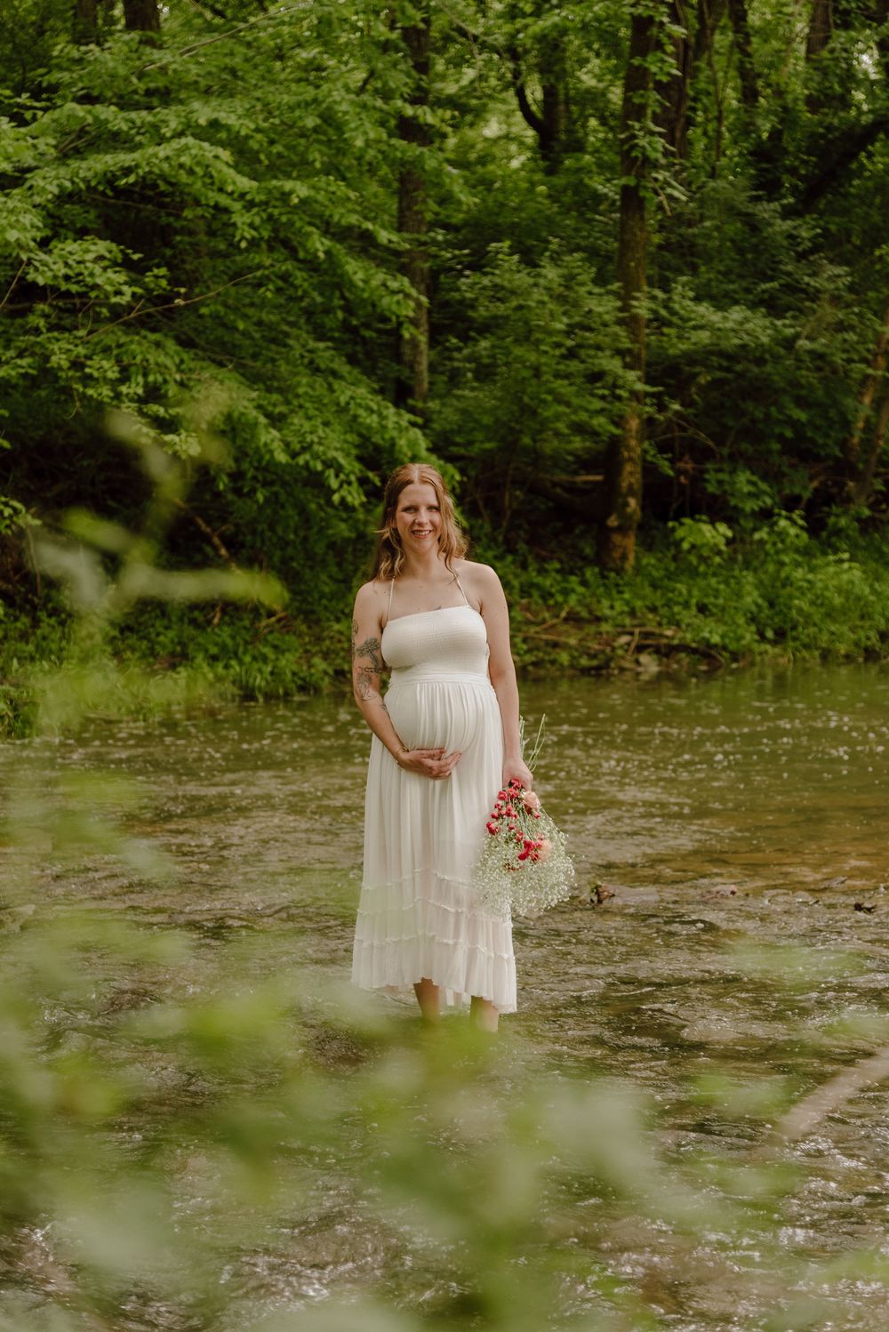 Pregnant woman in white dress holds flowers while standing in a shallow creek surrounded by lush green forest.