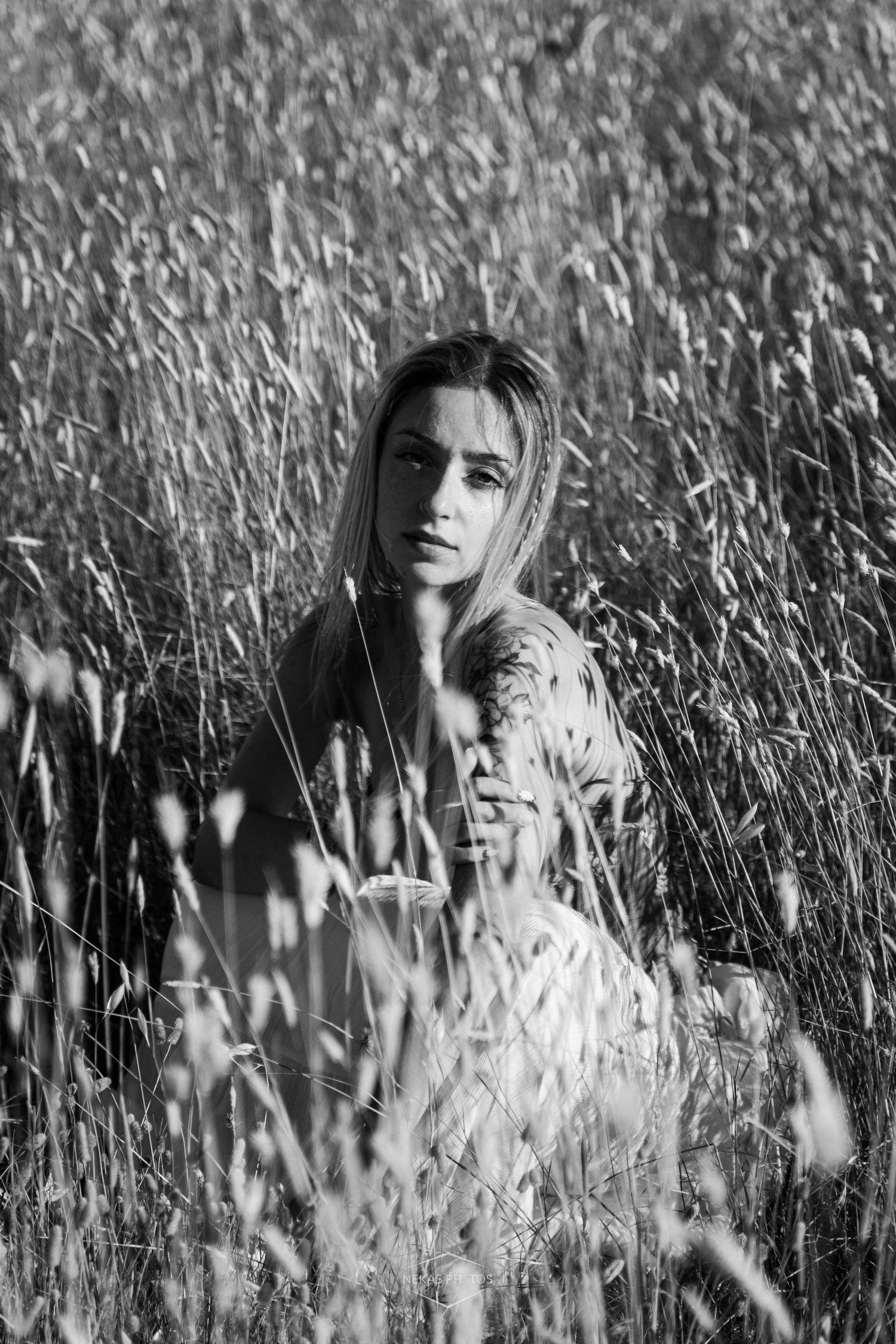 Black and white portrait of a young woman sitting in tall grass field, looking at camera with soft natural light.